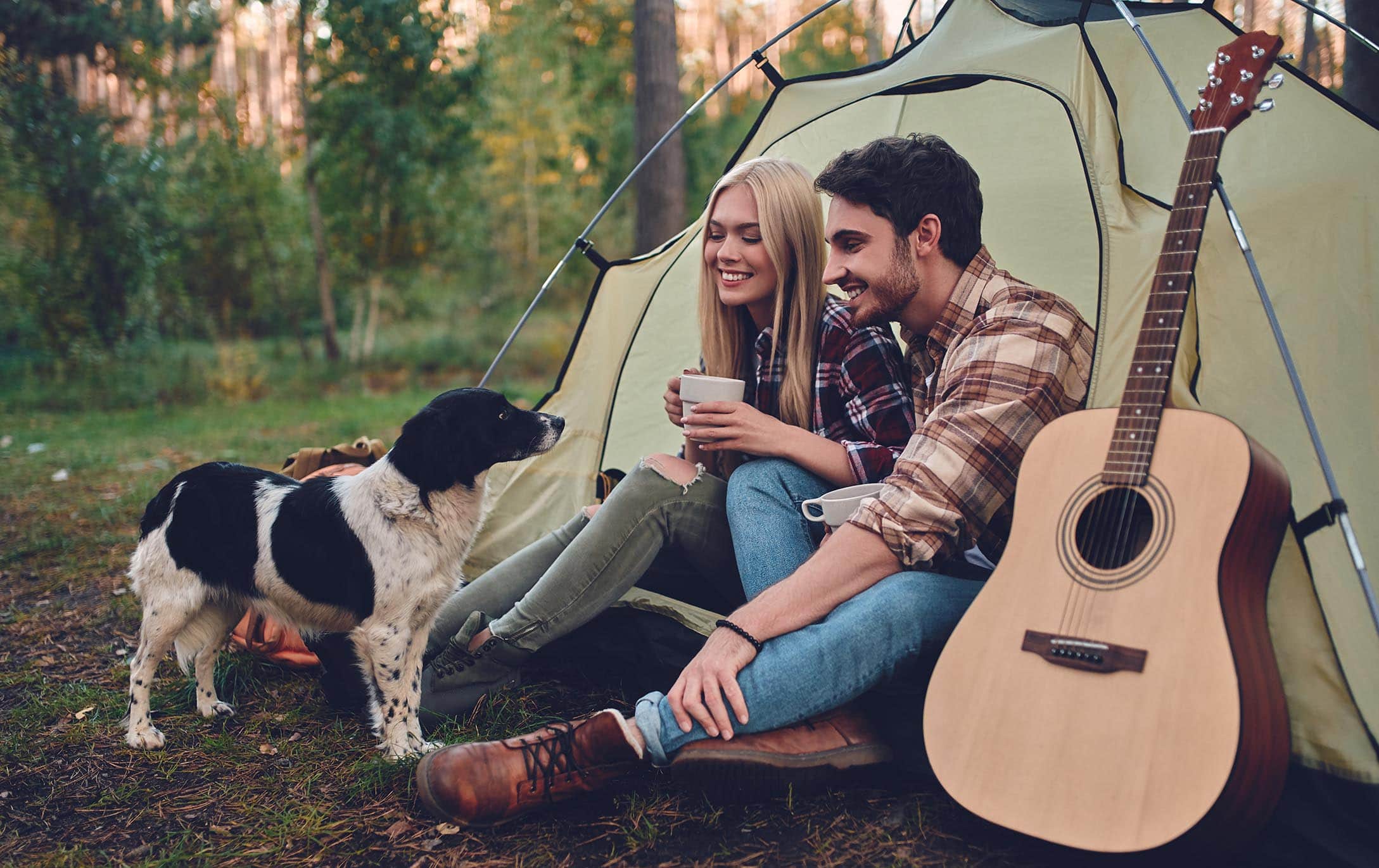 Image of smiling couple in front of tent with their border collie and an acoustic guitar.