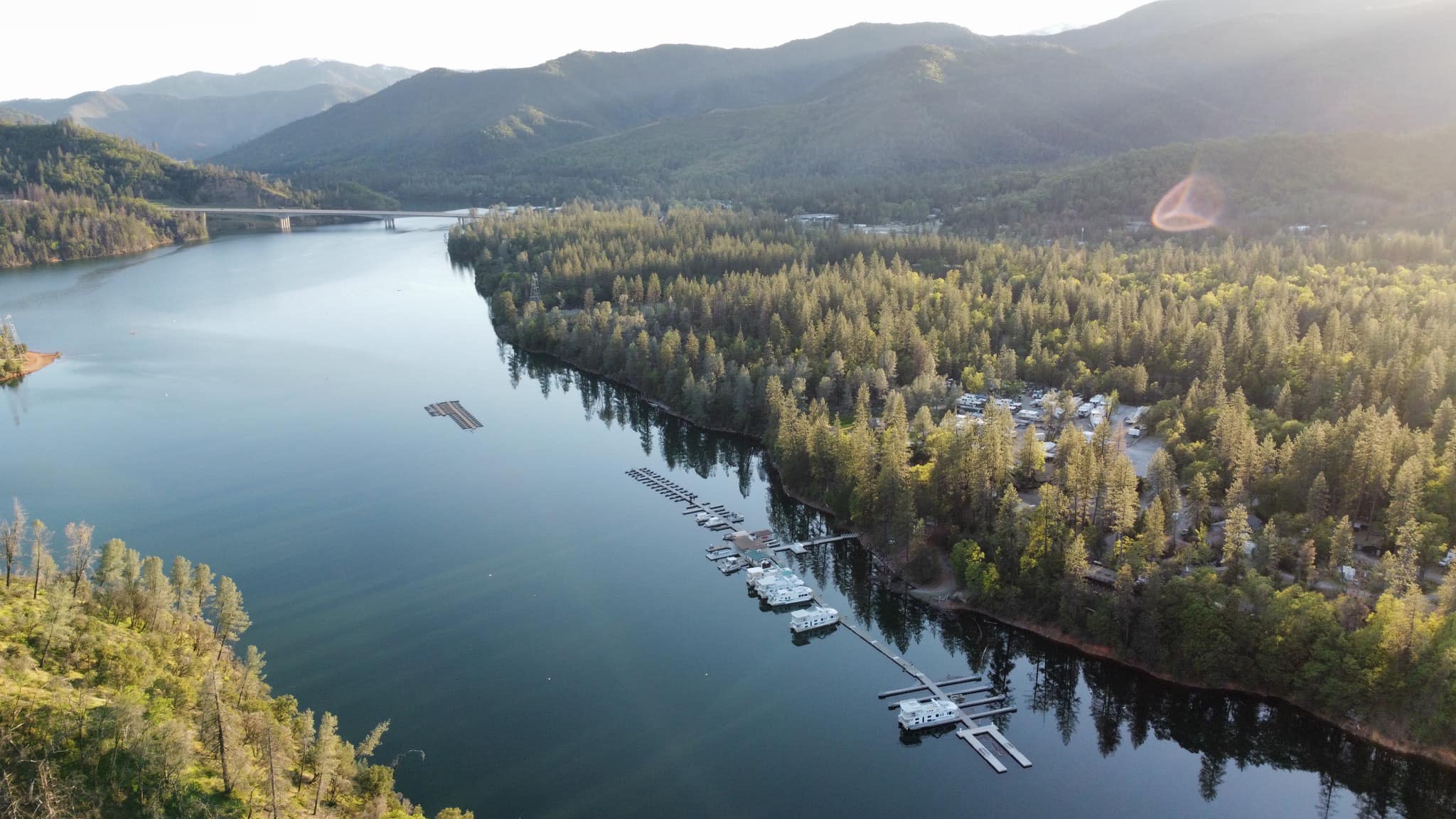 drone image of Antlers Resort and Marina shows the large body of water filled with boats of various sizes, from small rowboats to large houseboats. There are several banks along the lakes edges where people can dock their boats or sunbathe. The sky above is clear and blue. The large mountains provide a contrast against the lush green trees that line their base.